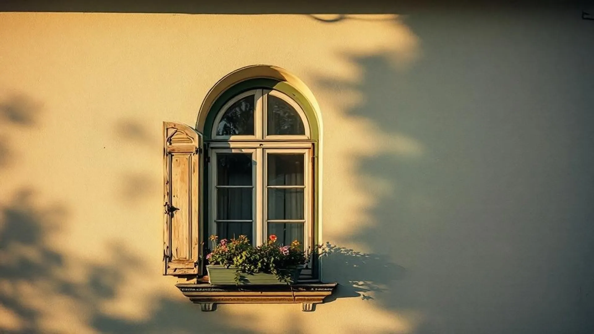 Detail einer süddeutschen Hausfassade: ein einzelnes Bogenfenster mit verwitterten Holzläden und kleinem Blumenkasten, warmes Spätnachmittagslicht streift die cremefarbene Putzwand.